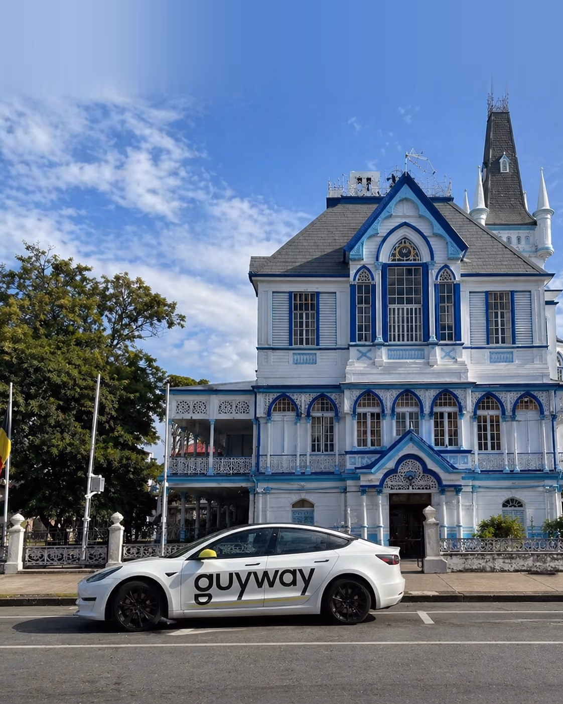 A GuyWay-branded electric Tesla Model 3 parked in front of Georgetown City Hall, the iconic blue and white Gothic Revival landmark in Guyana — running silently on Guyana's grid with zero tailpipe emissions