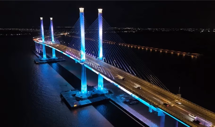 The Demerara Harbour Bridge lit in blue and violet at night, cables extending over dark water.