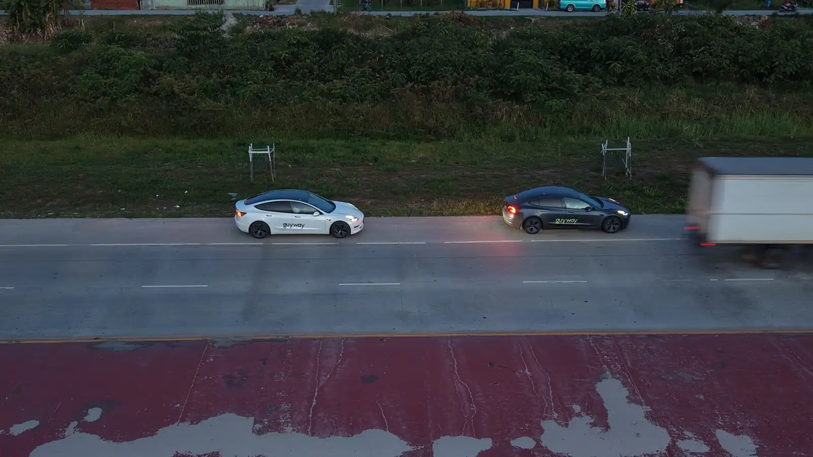 White and dark grey GuyWay Teslas driving along a concrete road beside a red median, seen from above