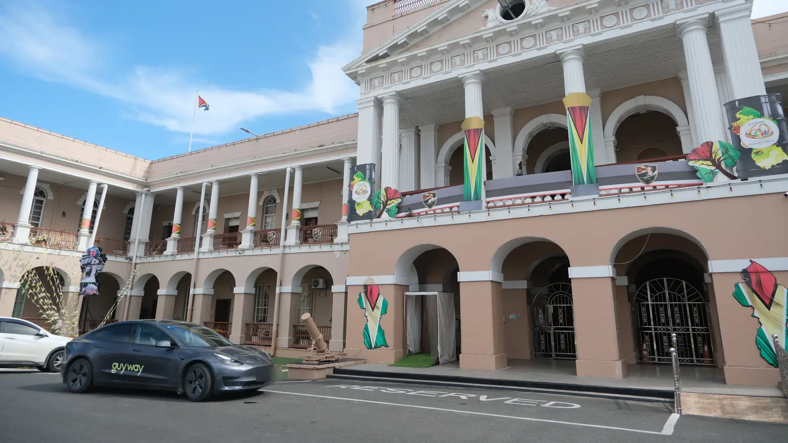 GuyWay Tesla Model 3 outside the Parliament Building in Georgetown, with Guyana flags on the facade