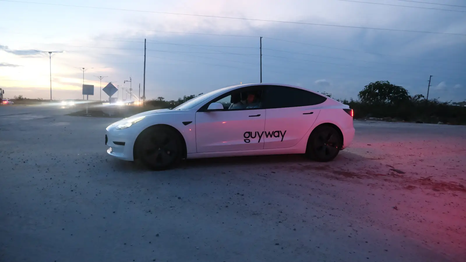 White GuyWay Tesla Model 3 with its headlights on at dusk, side view, power lines and road signs behind