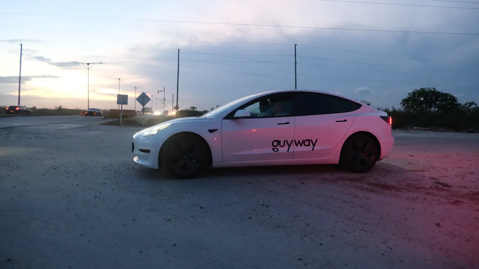 Tesla Model 3 blanco de GuyWay al atardecer, visto de lado, en un camino de grava