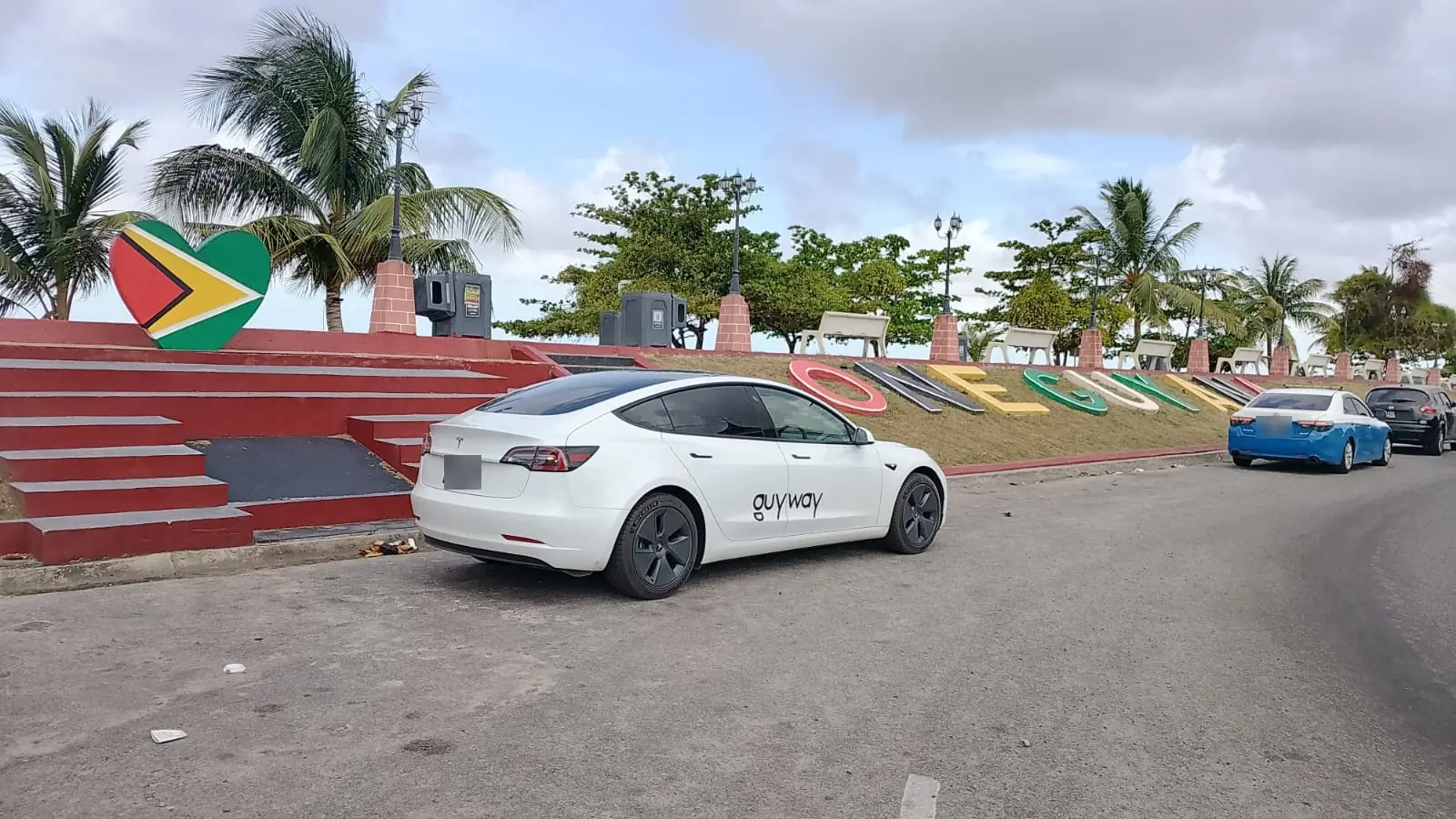 White GuyWay Tesla parked beside the One Guyana sign in Georgetown