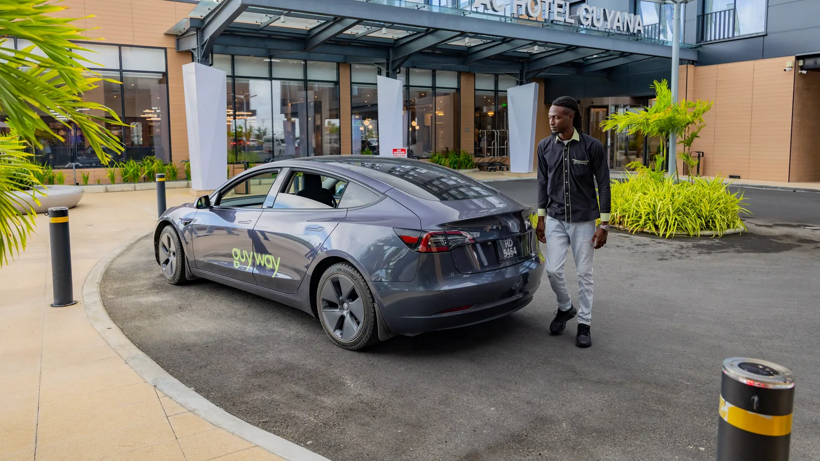 A GuyWay driver in uniform walks to a grey GuyWay Tesla at the entrance of AC Hotel Guyana