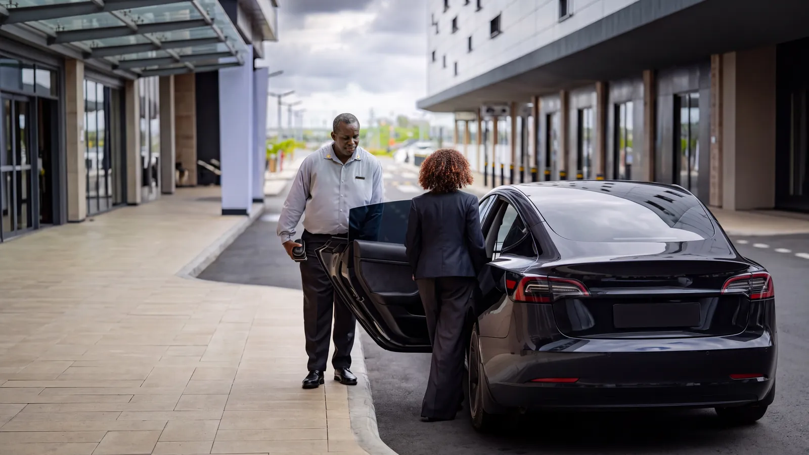 A GuyWay chauffeur holds the door of a black GuyWay Executive Tesla for a client at a hotel entrance