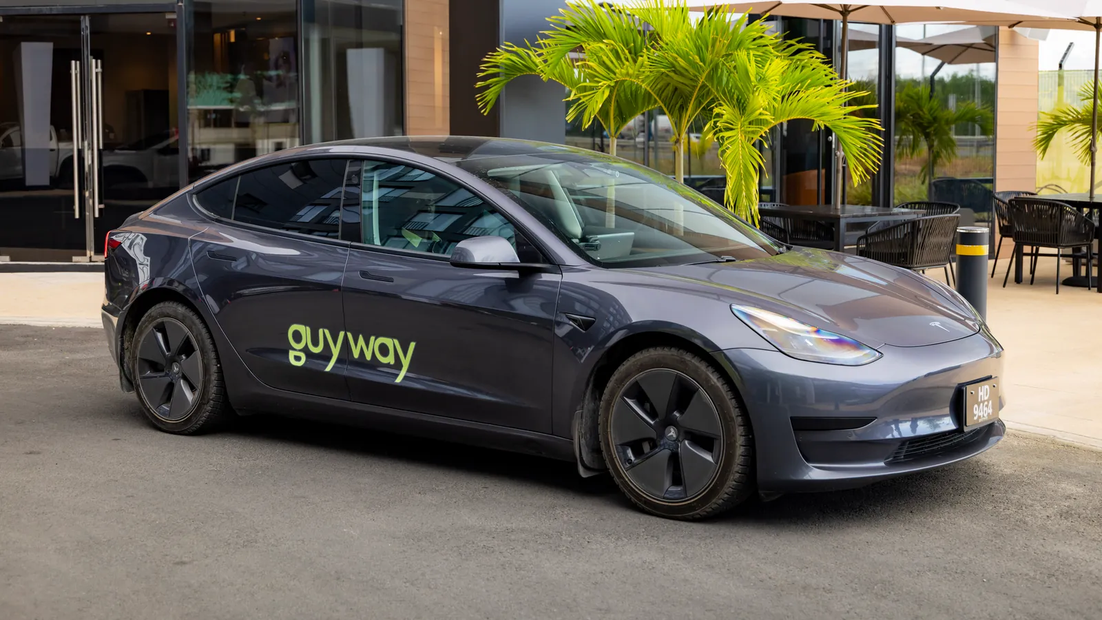Grey GuyWay Tesla Model 3 parked by the palm-lined terrace of AC Hotel Guyana