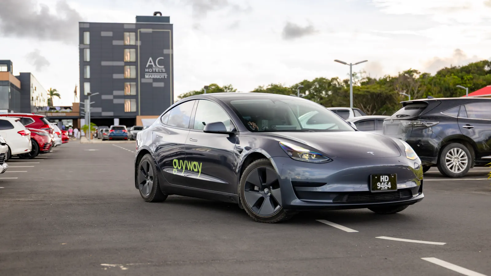 Grey GuyWay Tesla Model 3 in a car park, the AC Hotels by Marriott tower behind