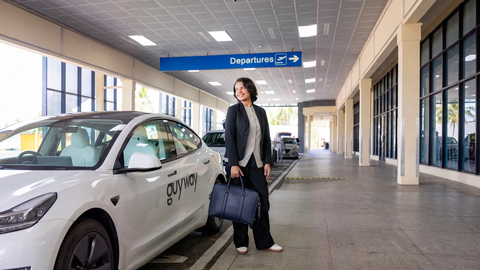 A smiling rider with her bag beside a white GuyWay Tesla at the departures kerb of Cheddi Jagan International Airport