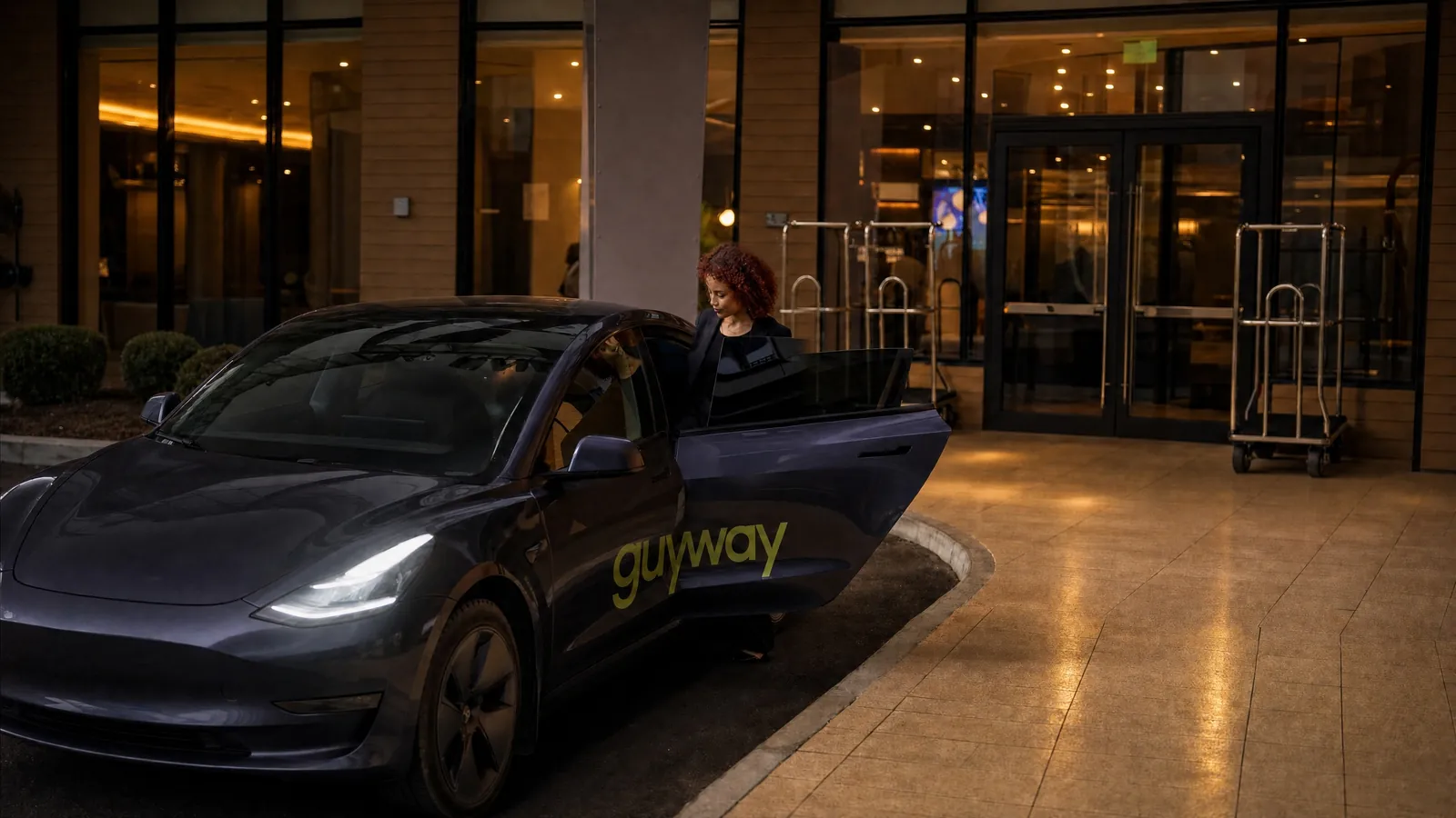 A rider gets into a grey GuyWay Tesla at a hotel entrance at night