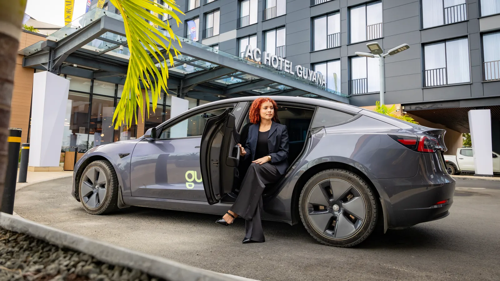 A rider steps out of a grey GuyWay Tesla under the entrance canopy of AC Hotel Guyana