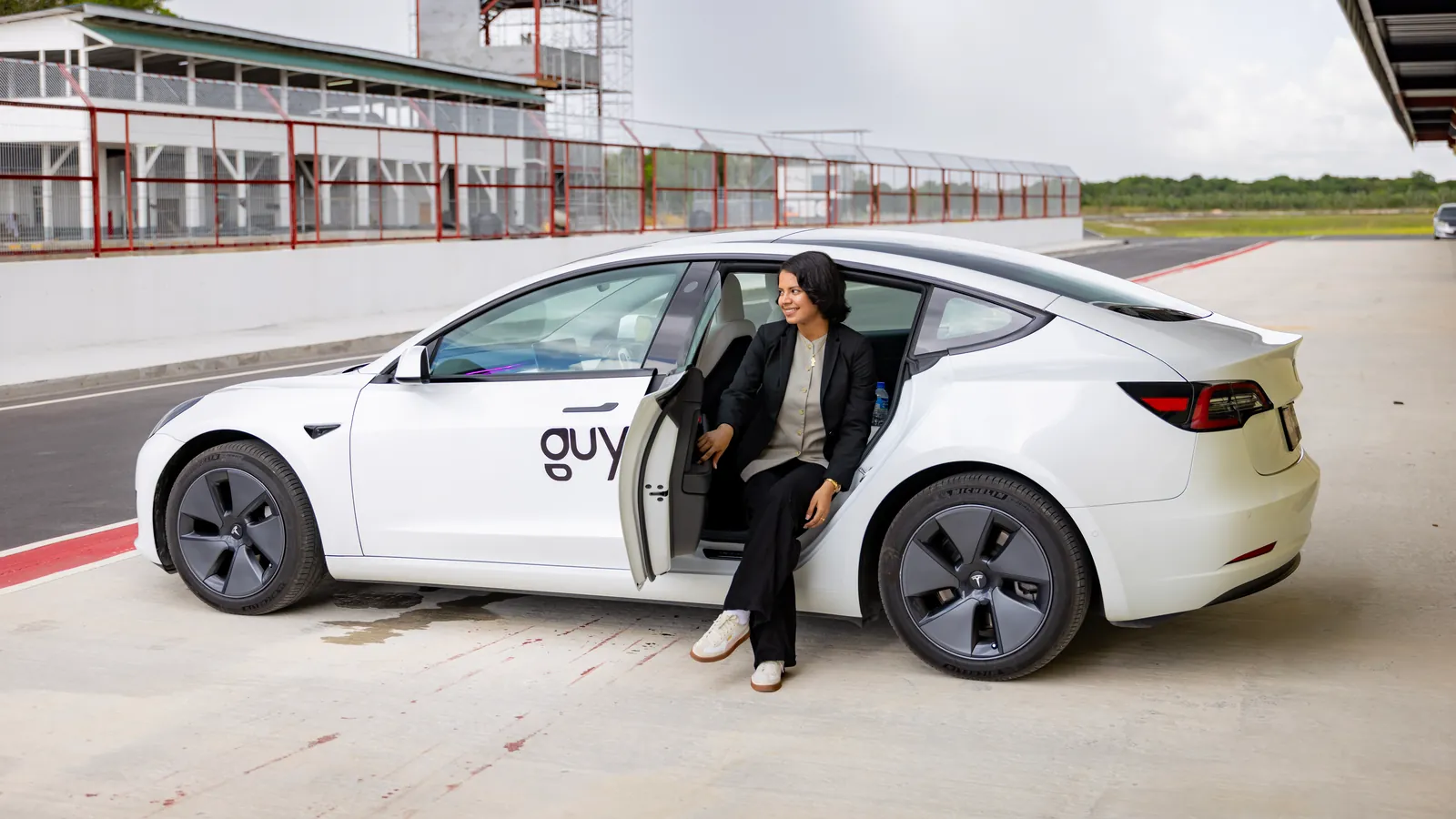 A rider sits in the open rear door of a white GuyWay Tesla in the pit lane of the South Dakota Circuit at Timehri