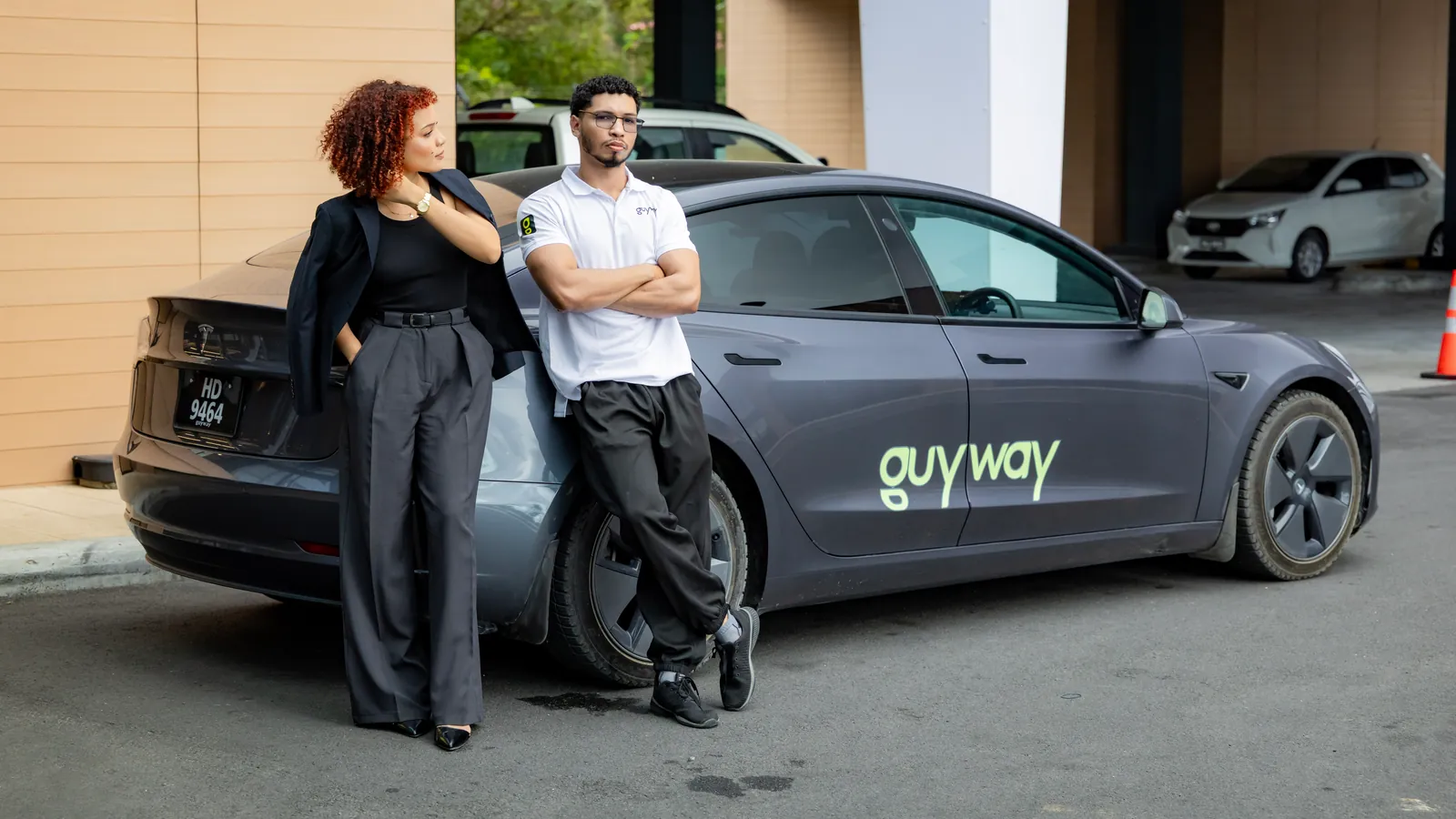 Two riders beside a grey GuyWay Tesla under a hotel entrance canopy
