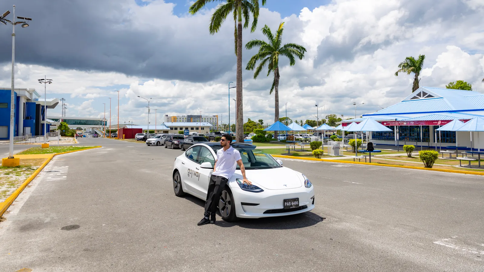 White GuyWay Tesla Model 3 in a palm-lined car park near Cheddi Jagan International Airport