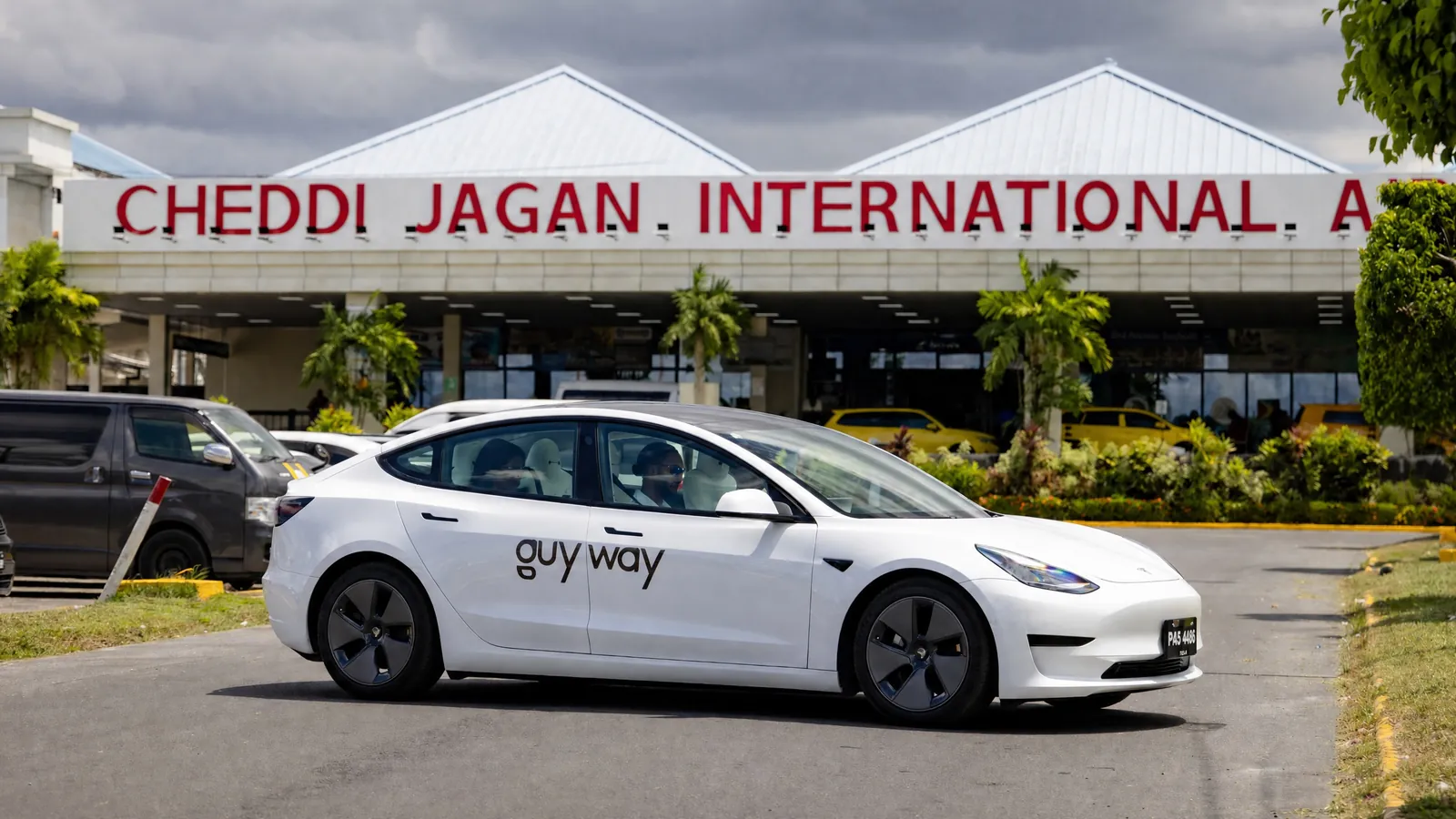 White GuyWay Tesla Model 3 driving past the Cheddi Jagan International Airport sign