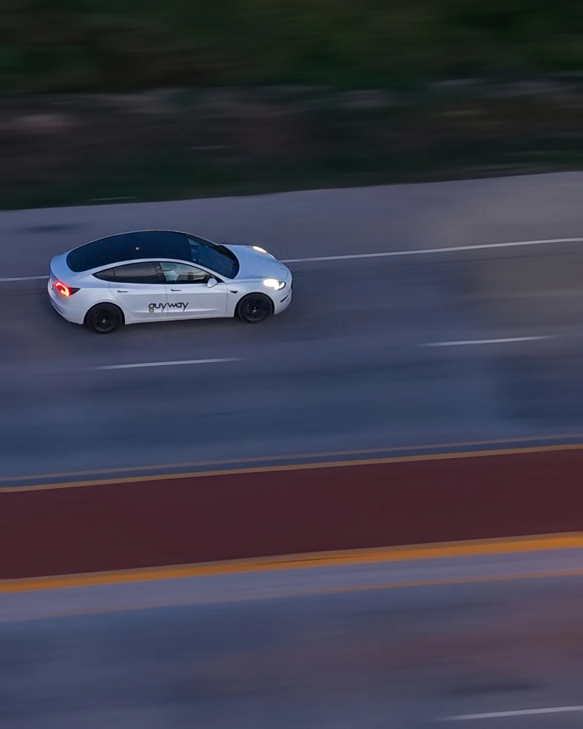 A white GuyWay Tesla Model 3 driving on the East Bank Highway at dusk, seen from a drone