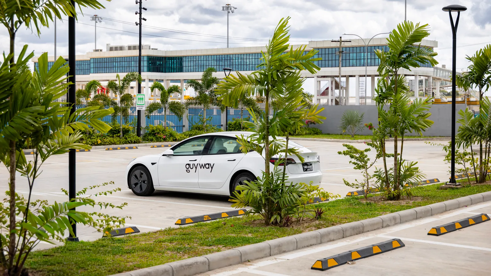 White GuyWay Tesla Model 3 in a palm-lined car park, the Cheddi Jagan International Airport terminal behind